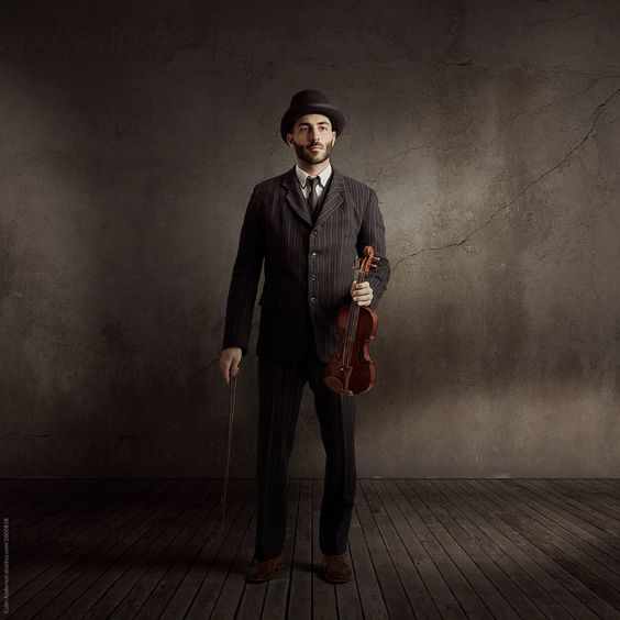 a man in a pinstripe suit and a bowler hat stands in a room with a violin and a cane in his hand. the room has dark walls and wood flooring.