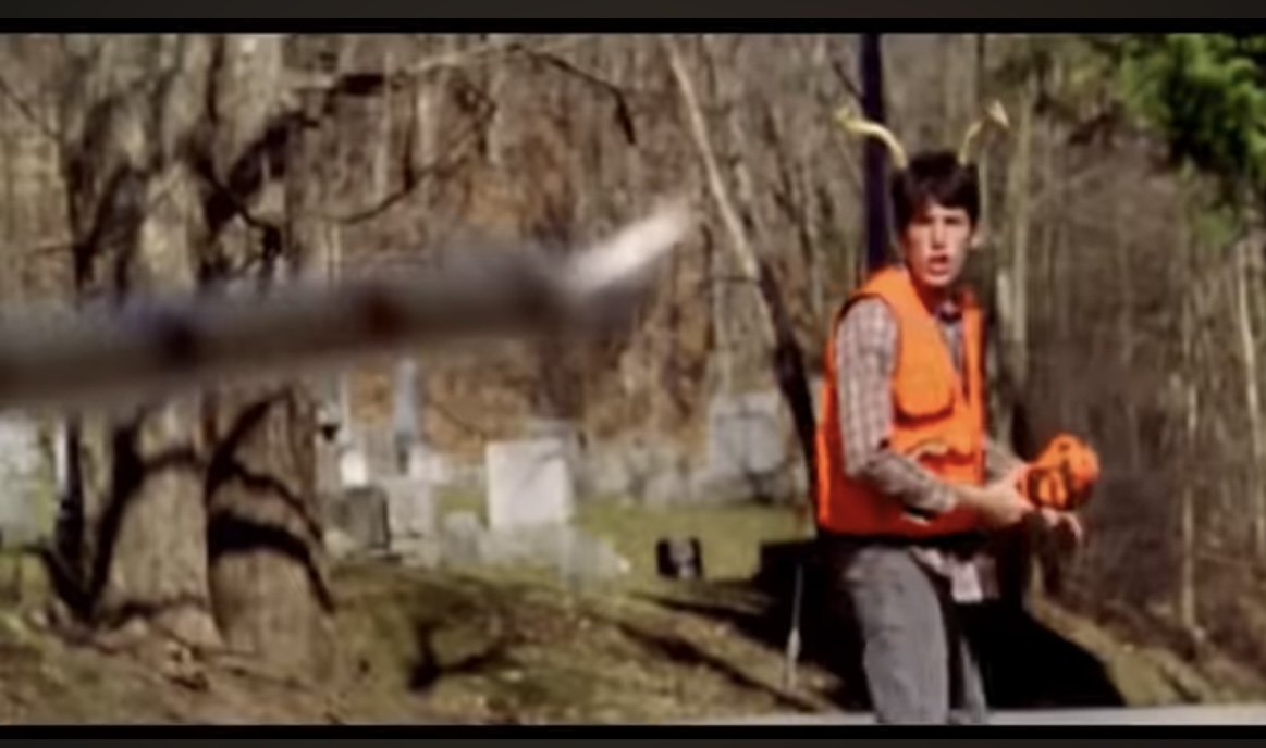 a man wearing an orange vest is in a forest holding a rifle, he appears to be looking at the camera but is blurry.  behind him are trees with no leaves and it looks to be autumn or fall.  the trees are bare and it looks to be cold.