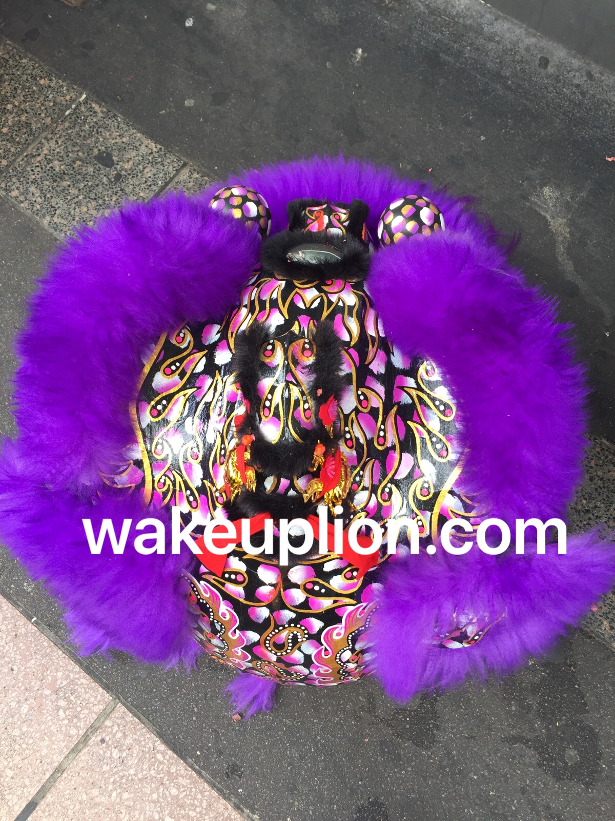 a purple lion dance costume with gold, black, and pink details is lying on a gray concrete surface. the costume is decorated with intricate patterns and has large, fluffy purple fur. the website "wakeuplion.com" is superimposed on the image.