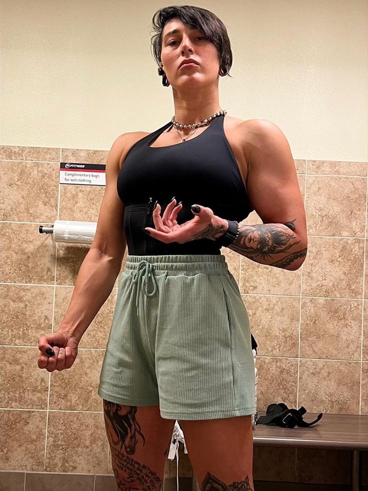 a woman with a muscular physique poses in a bathroom stall. she is wearing a black tank top and mint green shorts. she has tattoos on both arms and legs. she is looking at the camera with a confident expression. she is a bodybuilder and is displaying her strength and power.