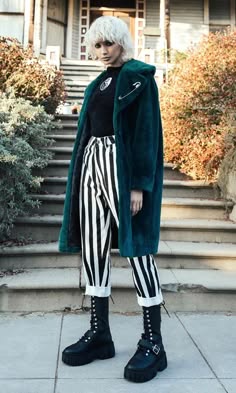 a blonde woman in a teal faux fur coat and black and white striped pants stands on the steps of a house.  she is wearing chunky black platform boots.  her hair is styled in a short bob, and she has a serious expression. she is looking down at the ground.  she is wearing a black turtle neck sweater with a small graphic on it.