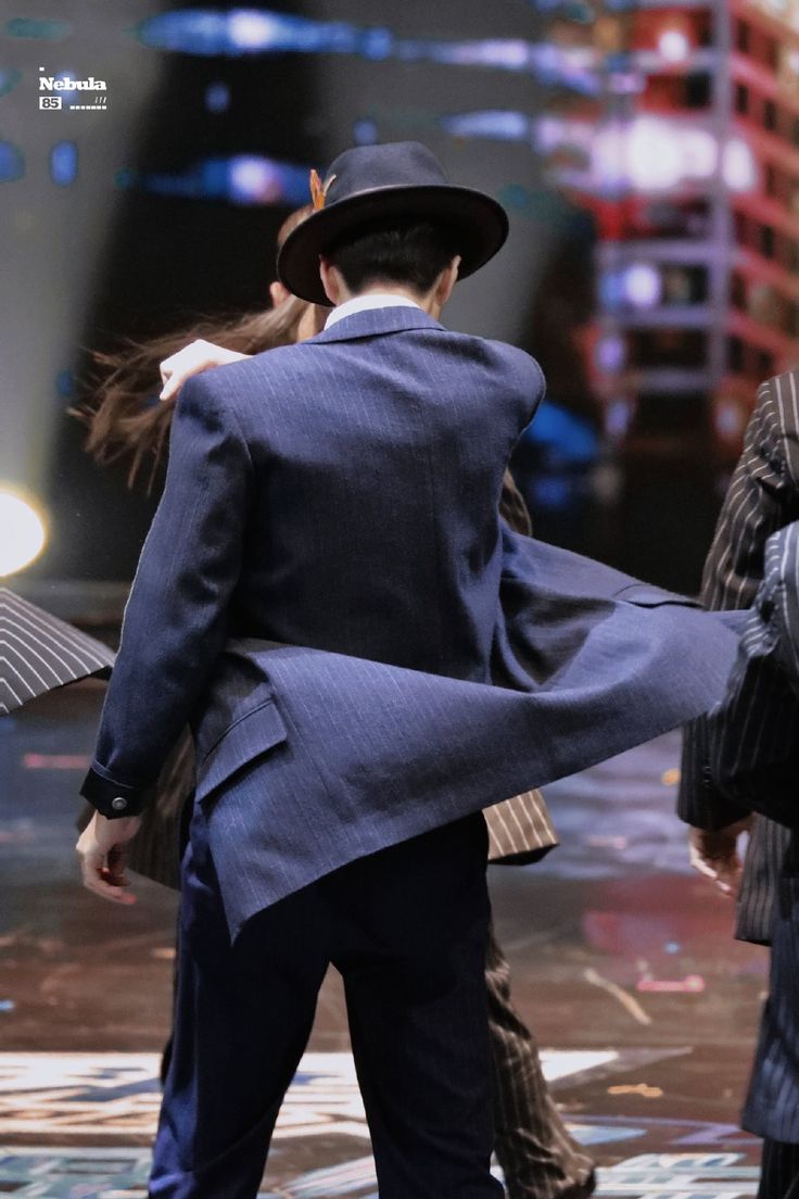 the image shows the back of a man in a dark blue pinstripe suit and fedora hat.  the wind is blowing his jacket. he appears to be walking and may be in a performance or event. the background is blurry, suggesting a city setting at night. another person in a similar suit is partially visible beside him. the overall impression is one of style, movement, and dynamism. the image has a slightly retro or vintage feel.
