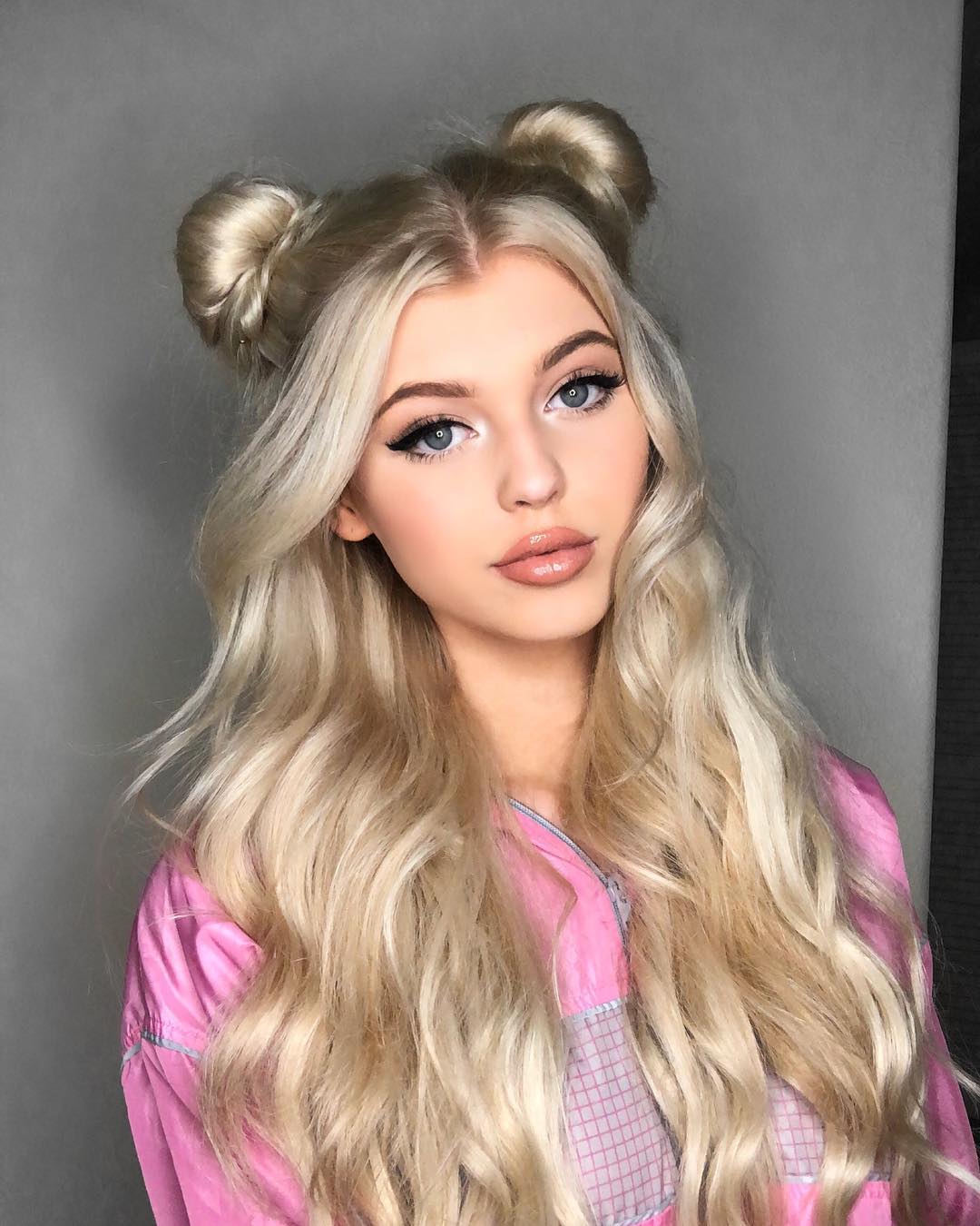 a portrait of a young woman with long blonde wavy hair styled with two space buns on top of her head. she has blue eyes, light skin, and is wearing makeup including eyeliner and lipstick. she is wearing a pink top. the background is a plain gray wall.