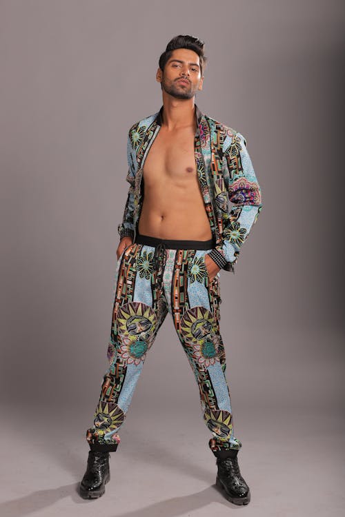 a fulllength studio shot features a male model dressed in a striking patterned ensemble against a neutral gray background. he is wearing a matching open shirt and pants set with a complex pattern of light blue with sun and geometric designs. the jacket is unbuttoned, revealing his bare chest. he is also wearing black boots. his hair is styled and he looks directly at the camera.