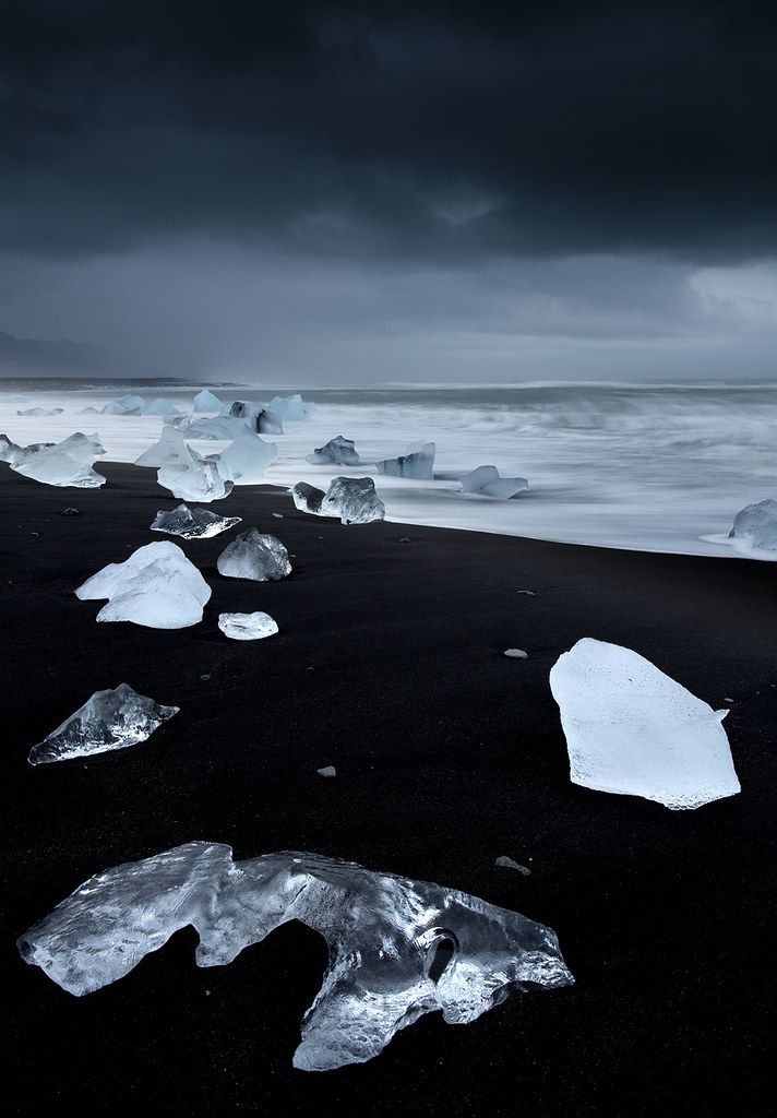 a dark and moody landscape shot of diamond beach in iceland, featuring icebergs scattered on the black sand beach, with the stormy sea and sky in the background. the waves are blurred due to a long exposure.