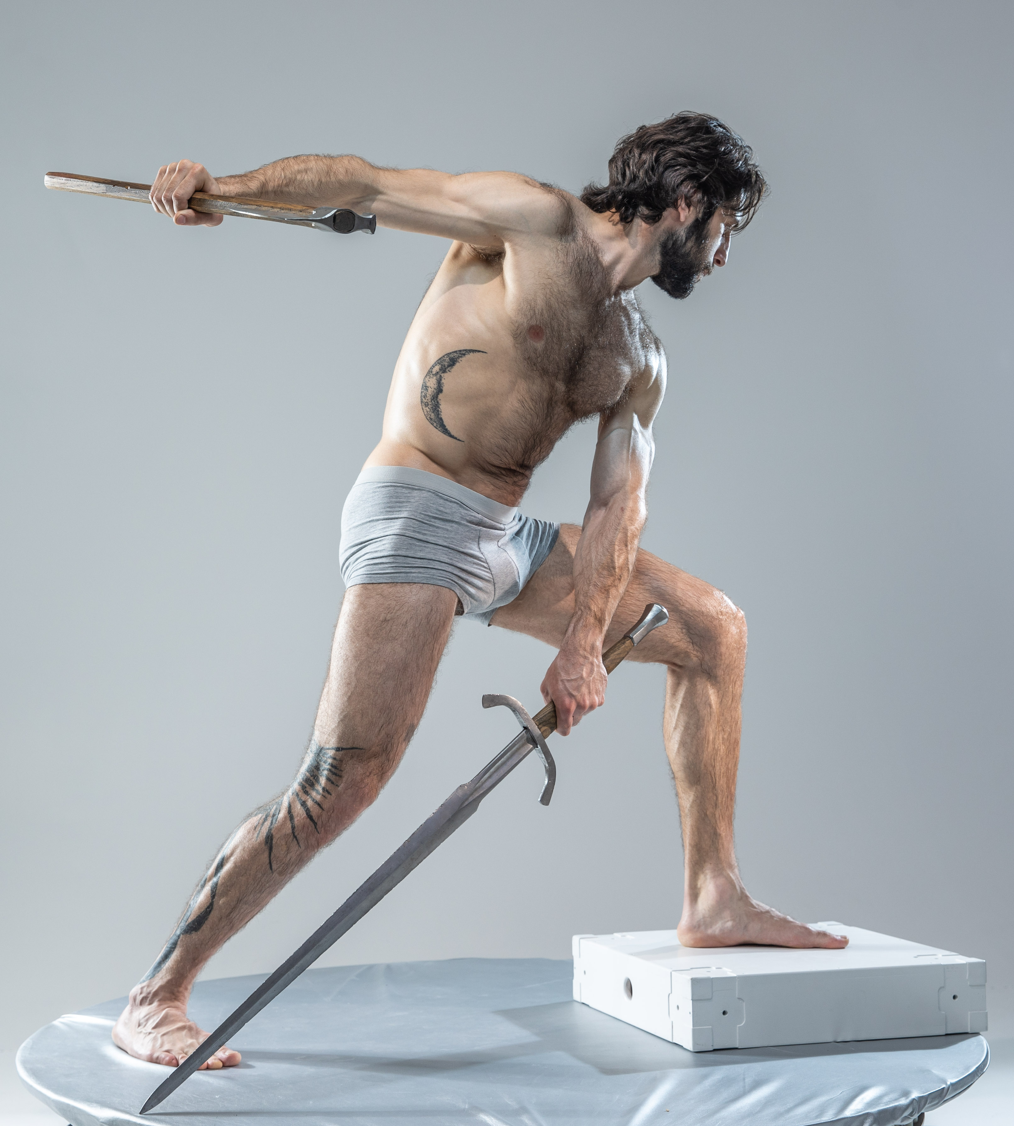 a muscular man with dark hair and beard  is captured in a dynamic pose against a solid grey background. he holds a sword in his right hand, ready to strike, and a short blade in his left hand. his body is angled, suggesting motion. he is wearing gray underwear and has tattoos on his left leg and right torso. the studio lighting highlights his physique and the intensity of the pose.  he stands on a round platform with a white block beside his right foot.