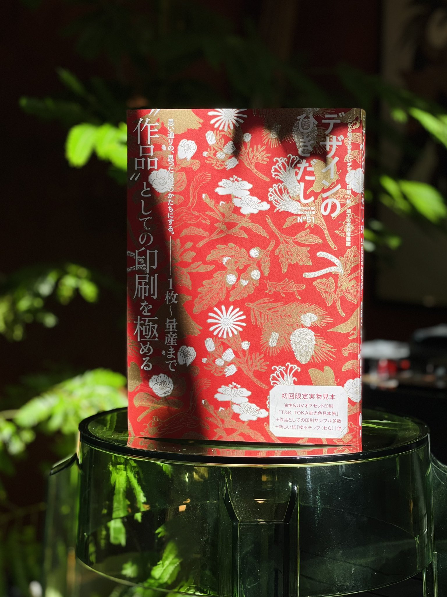 a red book with a floral design on the cover sits on a round glass table. the book is titled "デザインのひきたし" in japanese. the background is a blur of green plants, and the image is brightly lit with sunlight casting shadows.