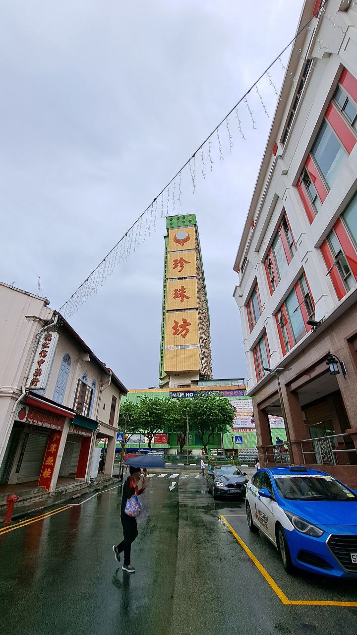 a wet street in a city, with a tall building with chinese characters on it in the background. there are shops on the left side of the street, and a car parked on the right side. a person is walking on the sidewalk. the sky is cloudy.