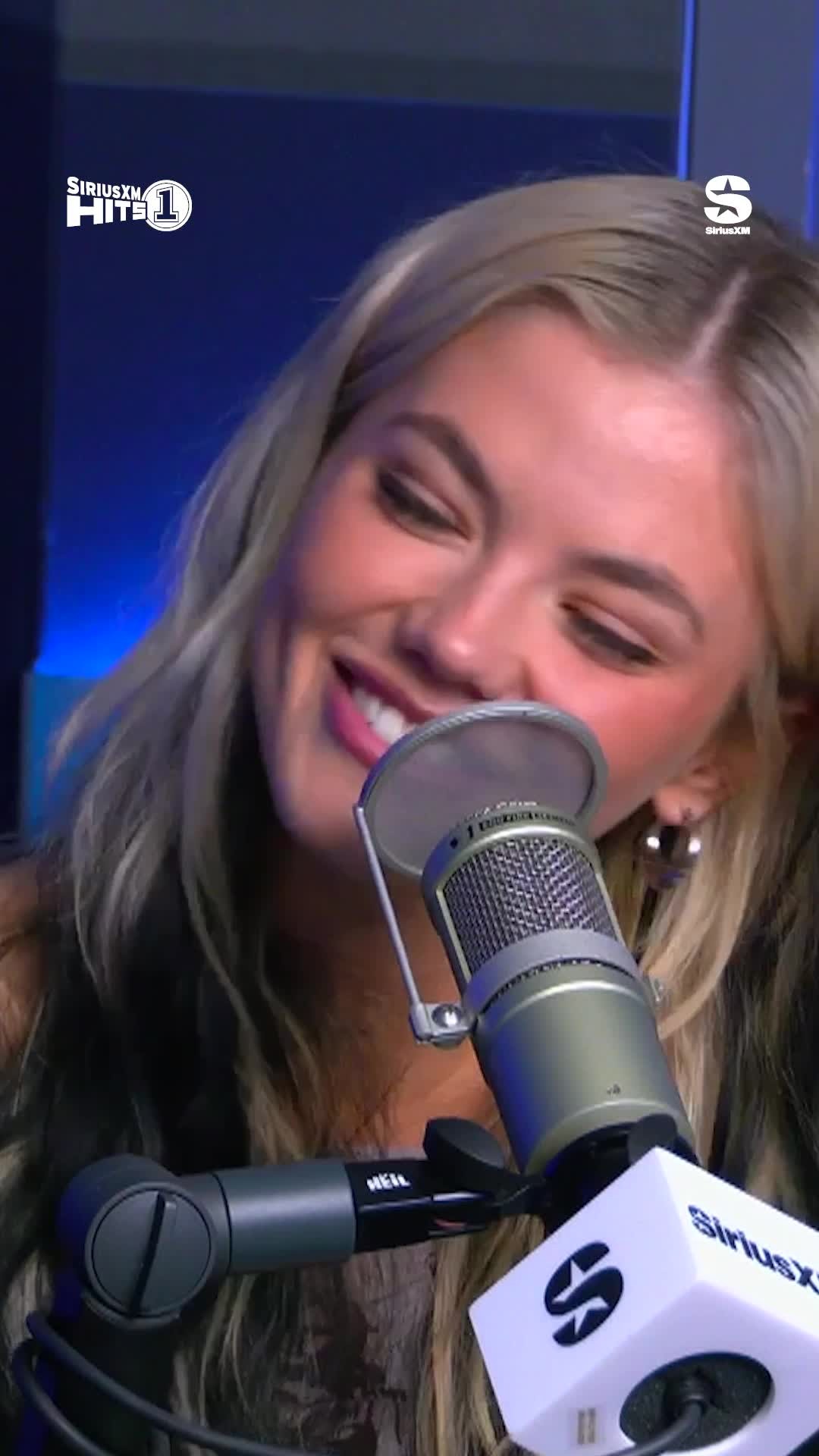 a closeup shot of a young woman with blonde hair and an earring, speaking into a microphone with the siriusxm logo on it. she is likely being interviewed in a radio studio. she smiles.