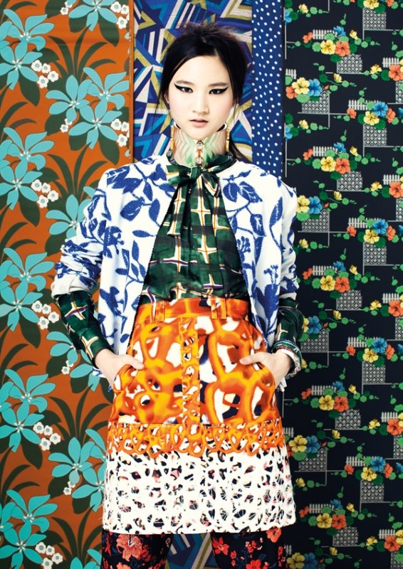 full shot of a stylish asian woman modeling a vibrant and eclectic outfit.  the model is positioned against a backdrop of various colorful floral and geometric patterned fabrics, creating a bold and textured visual contrast. she is wearing a white jacket with a blue floral pattern, a green and black plaid shirt underneath, an orange and white patterned skirt with a lower section of animal print, and dark patterned pants visible at the bottom. her makeup is striking, featuring defined eyeliner, and her dark hair is neatly styled. the overall image is a dynamic display of fashion and texture.