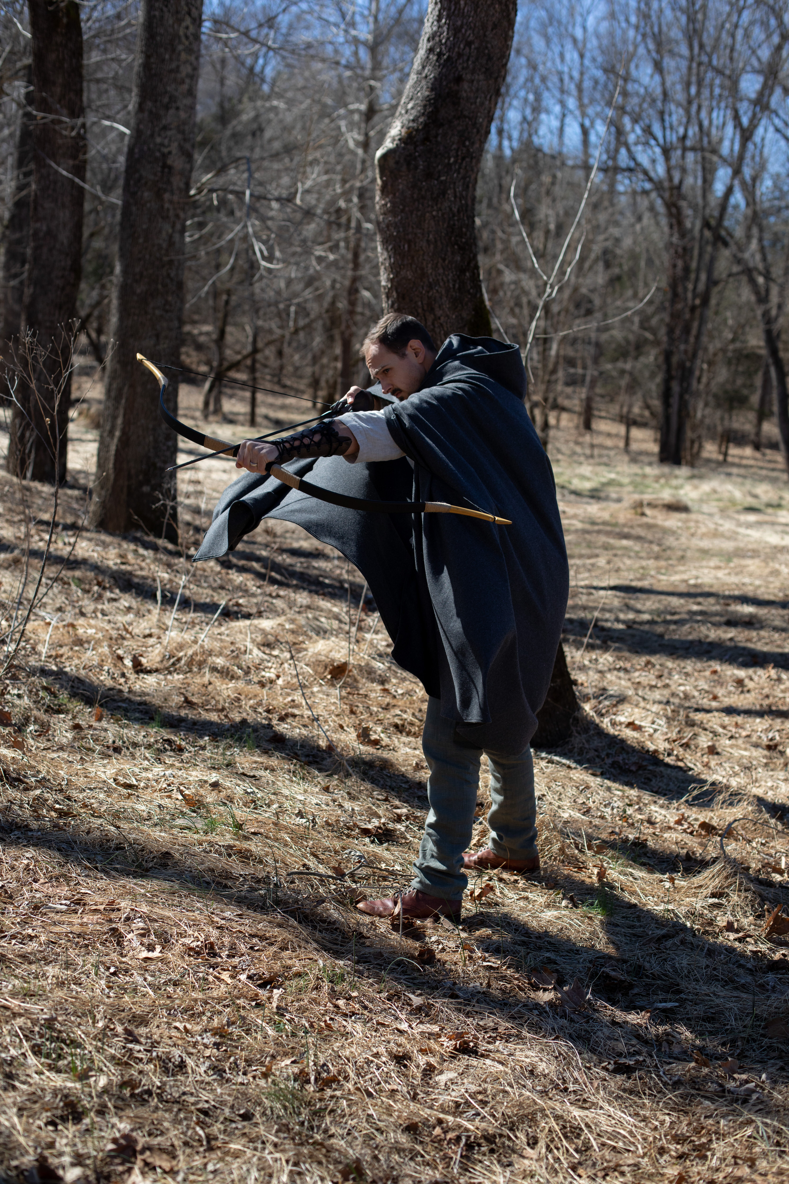 a man in a dark hooded cloak stands in profile in a forest, aiming a bow and arrow. he is focused on his target, and the arrow is about to be released. the scene is bathed in natural sunlight, with the trees casting long shadows on the ground. the overall mood is one of action and adventure.