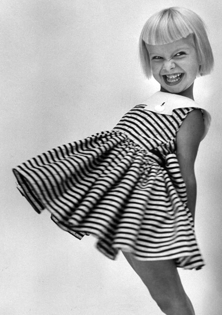 a black and white image shows a girl wearing a striped dress with a sailor collar, smiling widely. the dress has a full, flared skirt, and she is leaning slightly to the side, causing the skirt to billow out. her hair is short and styled with blunt bangs. the background is a plain, light color.