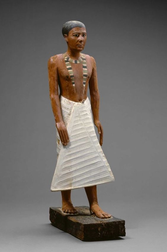 a fulllength shot of a painted wooden statue of an ancient egyptian man. the statue is standing on a rectangular base. the man is depicted with short black hair, brown skin, and is wearing a white pleated kilt. he is adorned with a beaded necklace.  the background is a neutral gray.