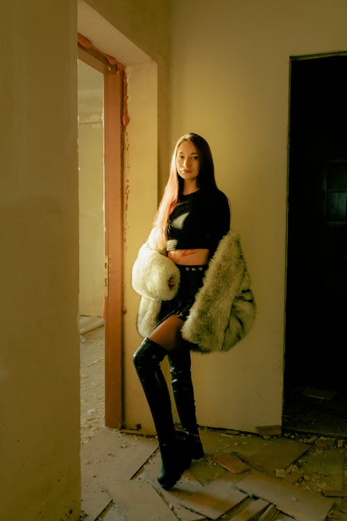 a young woman stands in a dilapidated building, wearing a black crop top and short pleated skirt, paired with a creamcolored faux fur coat and black thighhigh boots.  she is positioned near a doorway with sunlight streaming in, illuminating her and casting shadows. the background is a dimly lit, rundown interior with exposed walls and debris on the floor, creating a vintage or grunge aesthetic.  the overall style of the image is fashionable and slightly edgy.