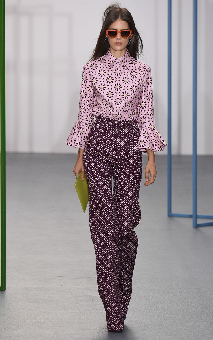 a full shot of a female fashion model walking the runway. she is wearing a pink and white patterned blouse with bell sleeves, paired with dark purple wideleg pants featuring a similar floral design. the model accessorizes with orange cateye sunglasses and carries a lime green clutch.  the background features a minimalist, lightgrey runway with light blue metal structures. the overall style evokes a retro or 70s vibe.