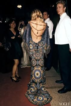 a group of people are posing for a photograph.  a woman in a stunning beaded gown is the center of attention. her dress has a vintage style and the intricate crochet design is eye catching. her look is complete with a necklace and earrings. the woman on the left is also beautifully dressed and poses with her arms crossed.  the man on the right is dressed in a tuxedo. he is smiling broadly. the trio is perfectly capturing the glamour and excitement of a formal event.  the background is blurred,  which gives a sense of depth to the image.  the scene is an event for a formal occassion.  the image is a classic example of elegant, formal fashion.