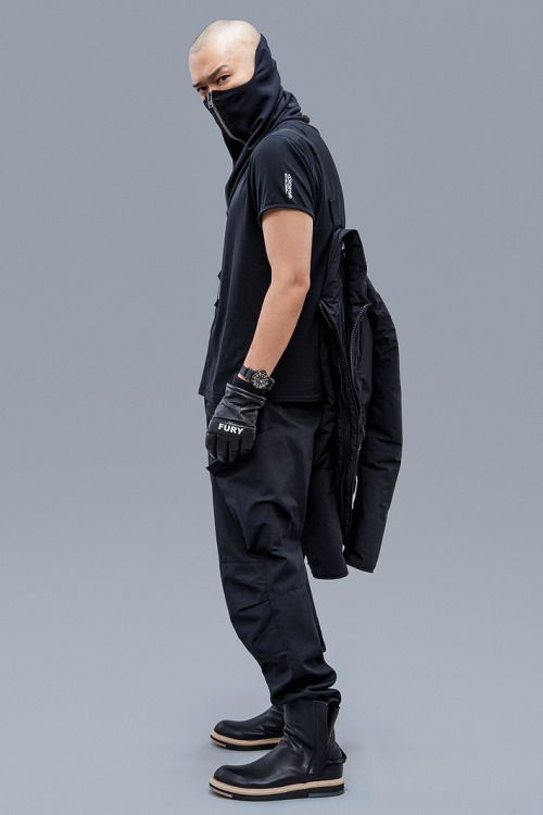 a man wearing all black clothing is standing with his right leg slightly bent.  he has a black balaclava covering his face, and he has on black gloves, pants, a jacket, and boots.  he is wearing a watch and a necklace.  the background is a light grey.