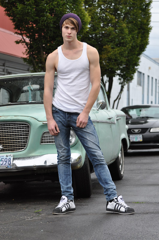 a young, attractive caucasian male model stands leaning against a vintage light teal car. he is wearing a white tank top, blue jeans, black and white adidas sneakers, and a purple beanie. the setting is outdoors during the day. the overall style is retro and cool.