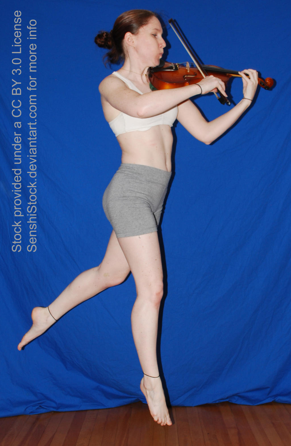 the image shows a young, athletic caucasian woman in a white sports bra and gray shorts dynamically playing a violin. she's midleap, legs extended, in a dynamic pose. the background is a solid blue, suggesting a studio setting. the overall style is indicative of a stock photograph, likely intended for use in various projects. the image includes a watermark indicating the license details.