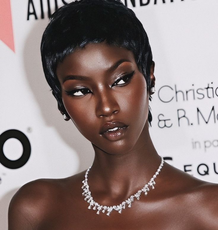 the image is a closeup portrait of a striking black woman with a short, stylish pixie cut. she has dark skin and is wearing elegant makeup, including prominent winged eyeliner. a sparkling diamond necklace adorns her neck.  her expression is serious and sophisticated. the background is a blurred white surface with partially visible text suggesting a highend event or awards ceremony.  the overall impression is one of fashion, beauty, and elegance.