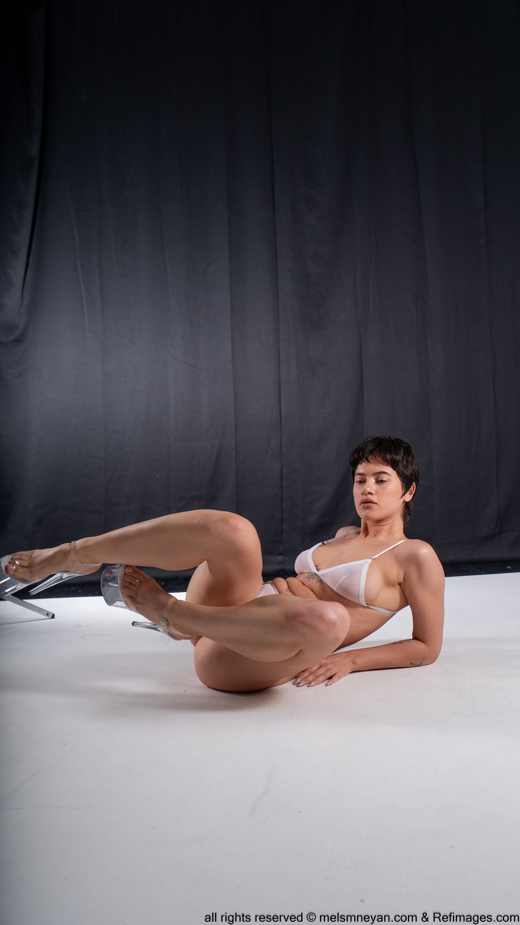 a full studio shot of a woman in white underwear and high heels lying down on a white surface in front of a black backdrop. she has short, dark hair. her legs are bent and angled to the left, and her right hand is resting on the floor. the woman is partially resting on a chair and her feet are in high heels.