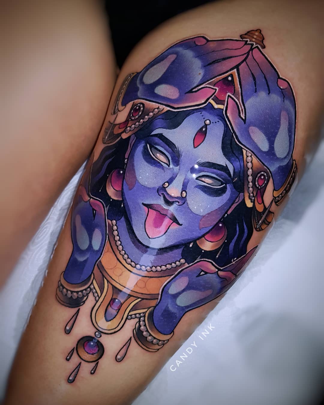 a closeup shot of a vibrant tattoo of the hindu goddess kali on someone's upper thigh. the tattoo is done in a neotraditional style with bold lines and shading, depicting kali with blue skin, a long tongue, and ornate jewelry.