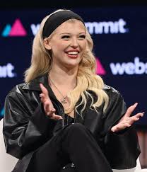 a woman with blonde hair wearing a black headband, a black jacket, and black pants is smiling and gesturing with her hands while speaking at an event. she is wearing a necklace and sitting on stage. the background includes a screen with the words "web summit" and other branding elements.