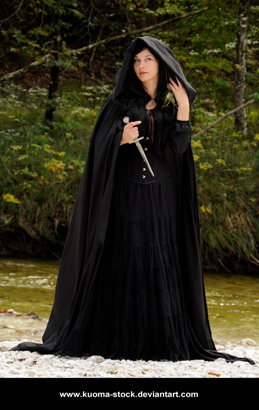a woman in a long, black hooded cloak stands by a river in a forest. she is holding a dagger and has a mysterious expression on her face. the scene is dark and atmospheric, creating a fantasy or gothic feel.  the woman's clothing is elegant and flowing, enhancing the overall mystical atmosphere. the setting appears to be autumn, judging by the colors of the foliage.