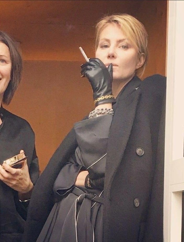 he image shows a closeup portrait of a stylish blonde woman in a black coat and black dress. she is wearing black leather gloves and several bracelets. she is holding a cigarette and smoking. another woman is partially visible in the background on the left side of the image. the background appears to be an indoor setting. the overall style is elegant and sophisticated.