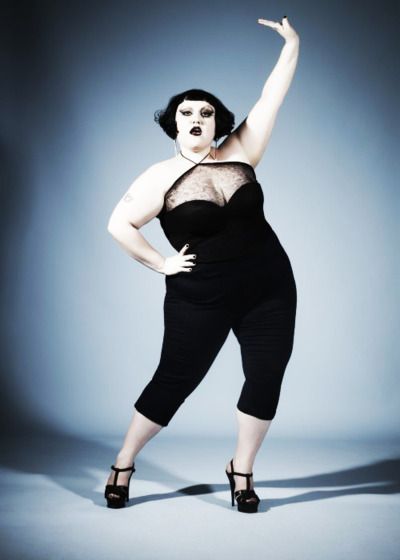 a fullbody studio shot of a plussize woman with dark bob hair and dark makeup. she is wearing a black jumpsuit with a sheer top and black high heels. she strikes a confident and stylish pose, her body angled and arm raised. the background is a muted bluegray.  the overall impression is one of elegance and selfassuredness within a context of bodypositive fashion.