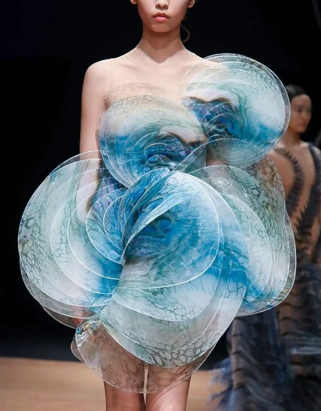 a fashion model is showcasing a beautiful, elaborate haute couture dress. the dress is made of  transparent layers of blue and white fabric, creating a stunning and intricate design. the layers are arranged in a way that resembles flower petals or overlapping circles. the model is standing on a runway, posing for the cameras. the background is blurred, highlighting the dress as the focal point.