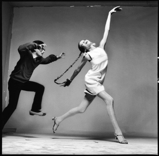 a black and white photograph of a woman in a white dress posing for a photo shoot. the woman is wearing a white dress and her hair is pulled back, giving her a classic and timeless look. she is posing in a graceful and artistic way, expressing the beauty and elegance of the fashion industry. the man on the left is the photographer, his face is only partially visible, he is facing left and holding a camera. this photograph captures a moment in time, a timeless piece of art that embodies the essence of fashion and beauty.