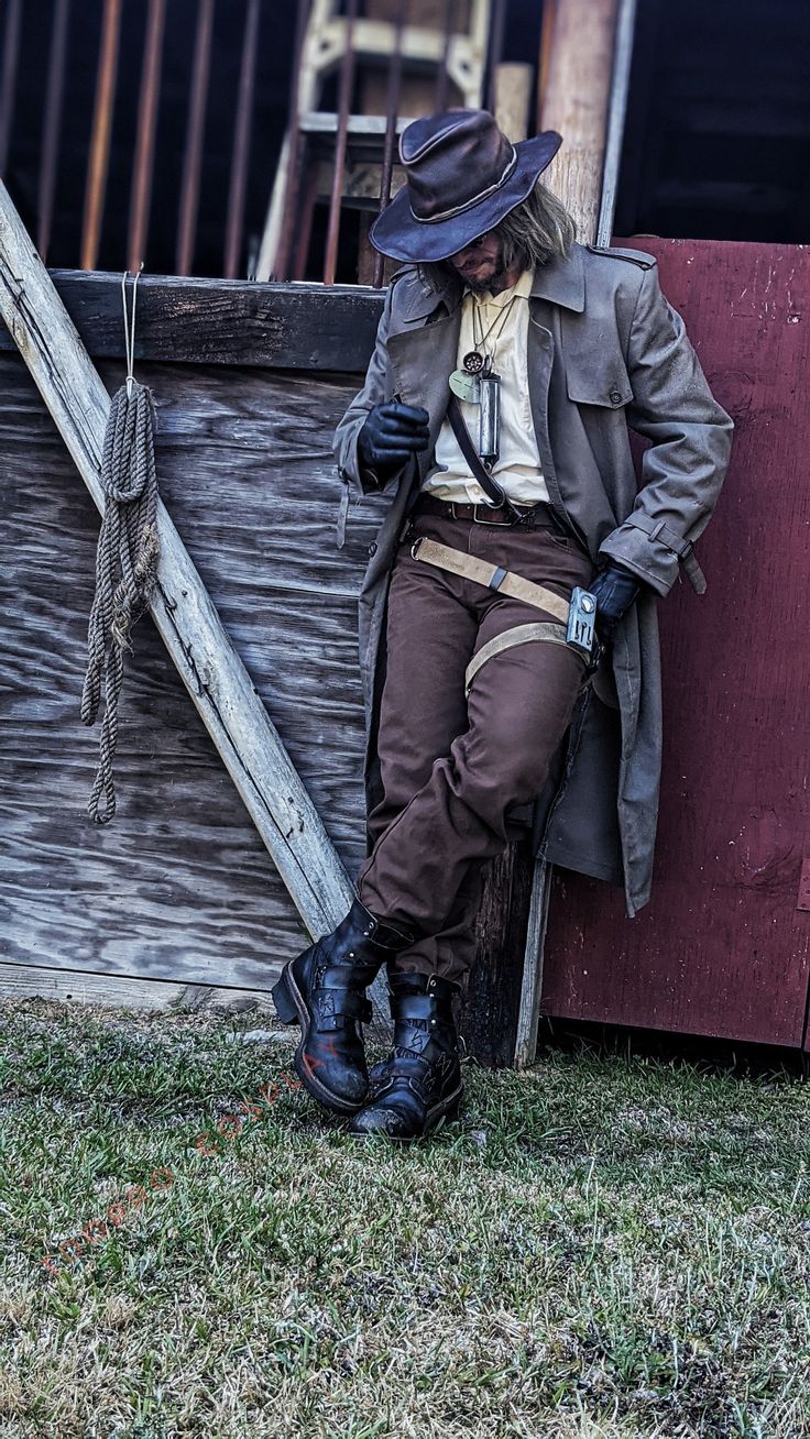 a full shot of a person in a detailed cowboy/steampunk cosplay costume, leaning against a weathered wooden structure. the individual is wearing a brown trench coat, brown pants, a hat, and sturdy boots.  they are holding a gun in a holster at their hip.  a rope hangs from the wooden structure behind them, and they appear to be lost in thought.  the overall tone of the image is moody and atmospheric. the setting appears to be outdoors in a grassy area.