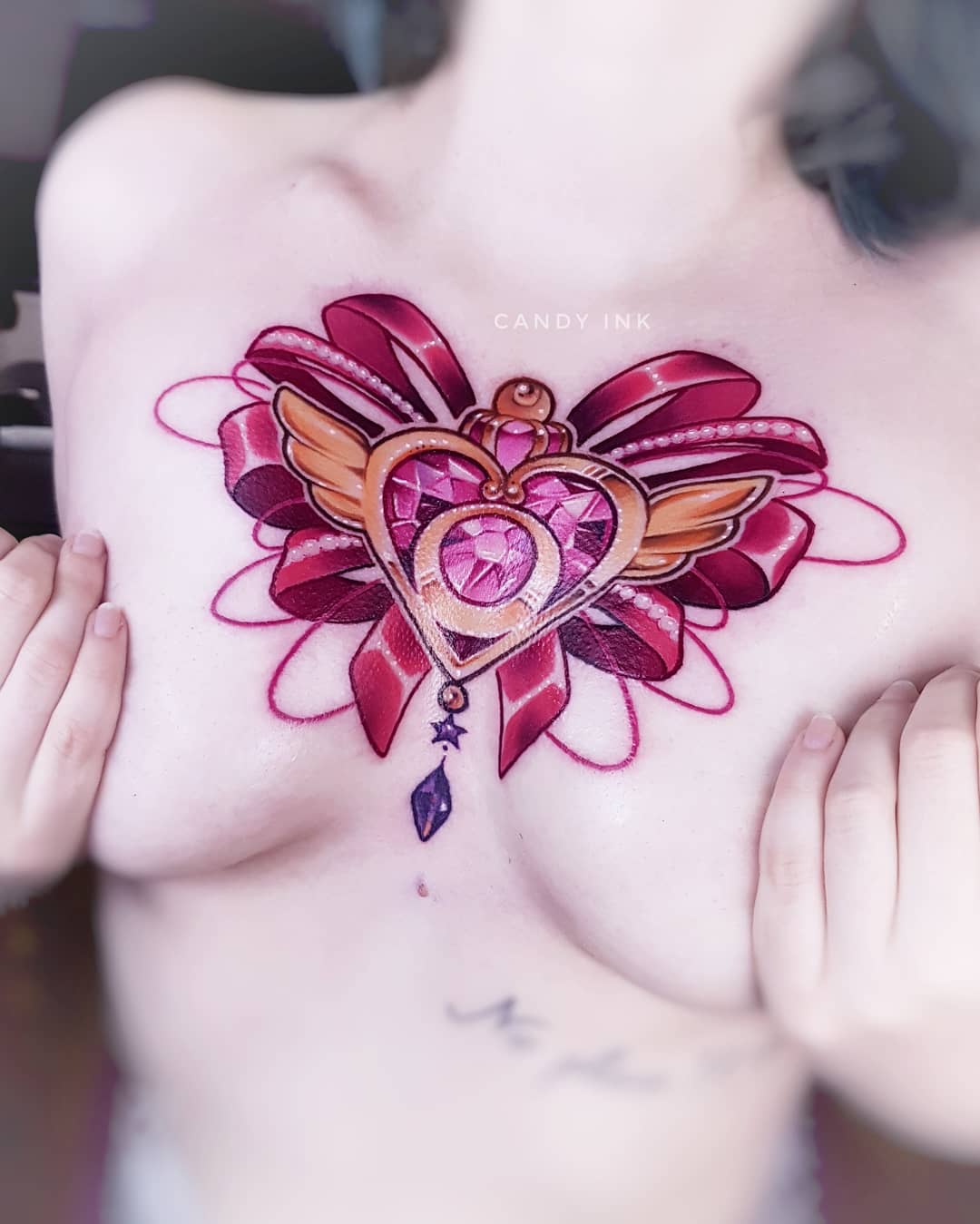 a closeup photo showcasing a vibrant sailor mooninspired tattoo inked on a woman's chest. the tattoo features the iconic crisis moon compact, rendered in a colorful and detailed style, with a large pink bow at its top. the woman's hands gently frame the tattoo, emphasizing its placement and intricate design. the lighting is bright, highlighting the tattoo's colors and the smooth texture of the skin.