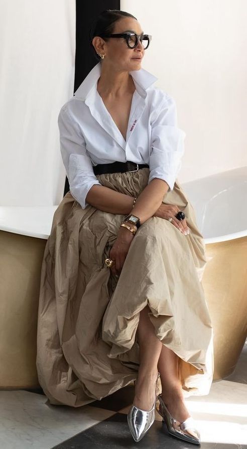 a stylish woman in a white shirt and brown skirt poses on the edge of a white bathtub.  she is wearing silver heels and has a watch and a ring on her left hand.  the woman is looking to the side and has glasses on her face.