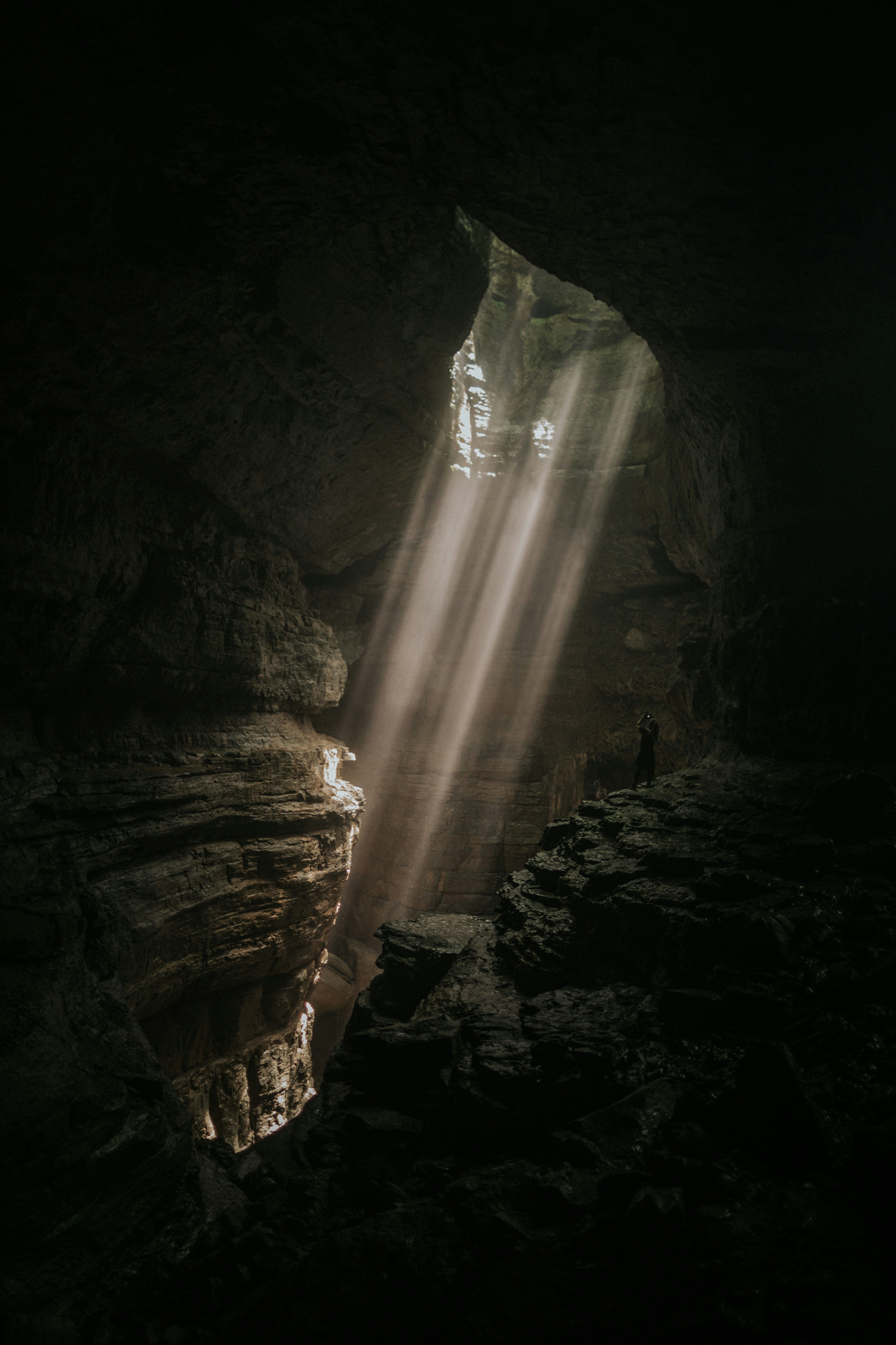 this is an image of a large, dark cave with a single opening at the top.  sunlight streams through the opening, illuminating dust particles in the air and creating dramatic beams of light. a lone figure stands on a rock ledge within the cave, silhouetted against the light. the image evokes a sense of awe, mystery, and the power of nature.