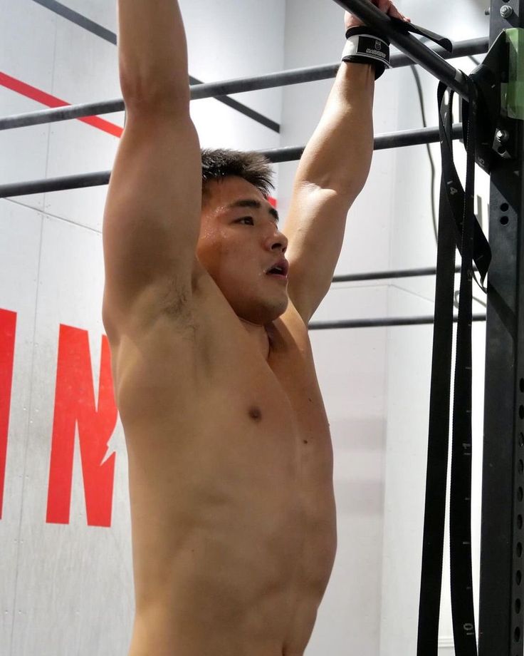 a man in a gym, hanging from a pullup bar, demonstrating a pullup exercise. he is shirtless and showing off his muscles. he is dedicated to fitness and is working towards his fitness goals.