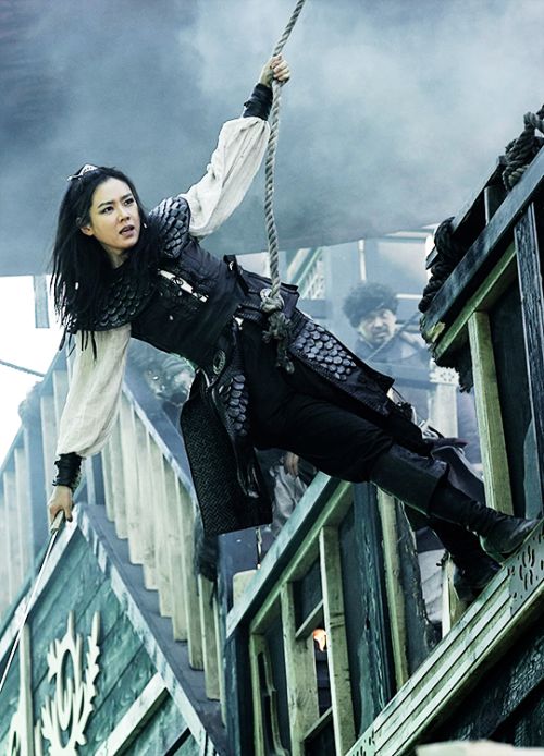 a woman dressed as a pirate is climbing a rope, she is holding a sword in her hand, and is looking towards the camera. she is dressed in a black and white costume, with a black band around her hair. she is on a ship that appears to be damaged.
