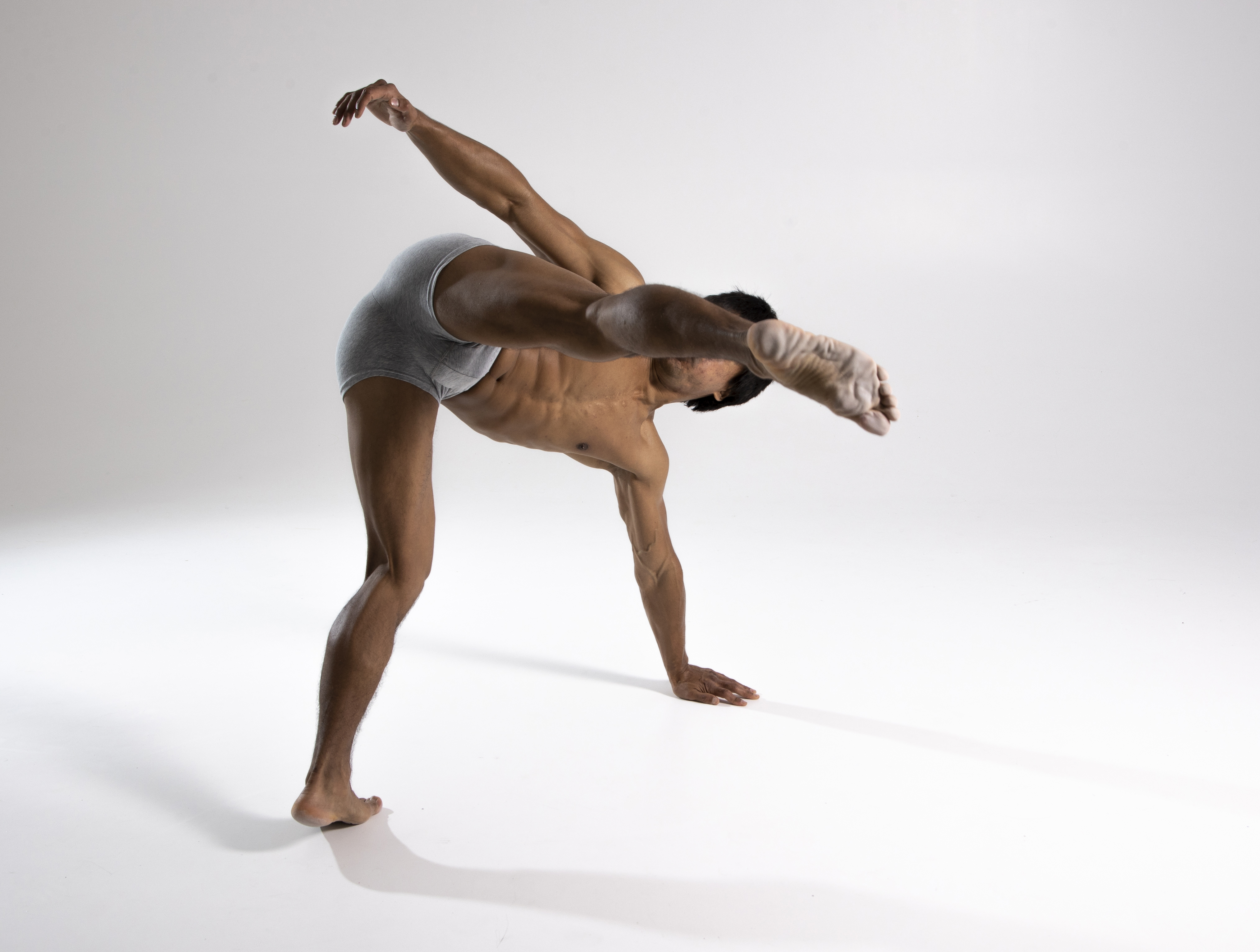 a muscular man is performing a contemporary dance move in a studio setting. he is wearing gray briefs and is barefoot. he is bent over with one hand and one foot on the ground, balancing, while his other arm is raised and the other leg is extended out to the side. the background is white with a subtle shadow. the dancer appears athletic and flexible.