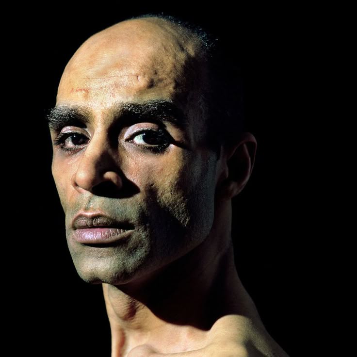 a closeup portrait of a bald man with a serious expression against a dark background. he has strong features, dark eyes, and is wearing makeup around his eyes. the lighting is dramatic, casting strong shadows across his face and body.