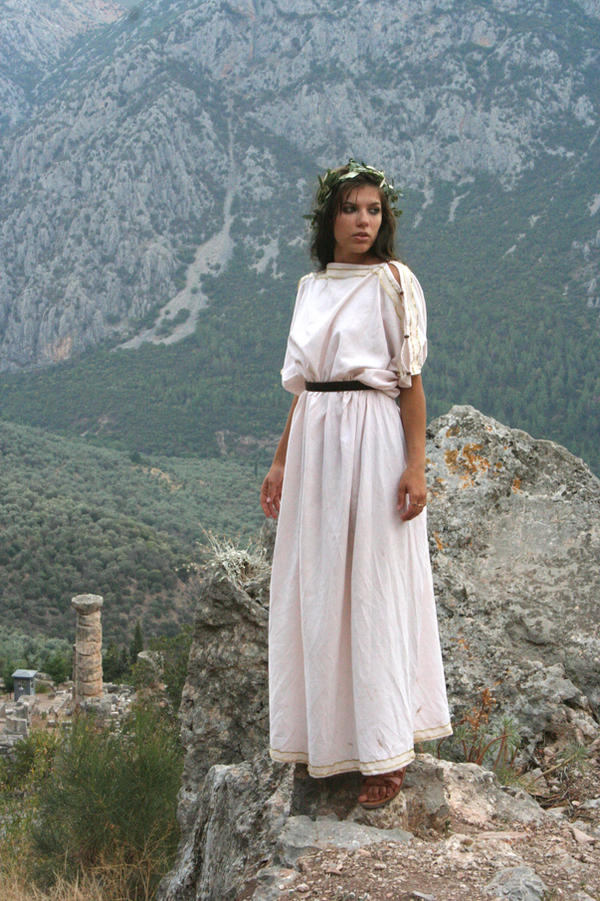 a young woman wearing a light pink ancient greekstyle chiton stands on a rocky outcrop overlooking a mountainous landscape. the ruins of an ancient structure are visible in the background.  she is wearing a leaf wreath and has a serene expression. the scene evokes a sense of history and ancient culture.