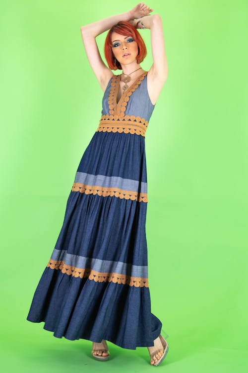 a fulllength shot of a woman with red hair wearing a blue denimlike maxi dress with brown trim, standing against a bright green backdrop. she is also wearing sandals and a necklace. she has her hands raised above her head.