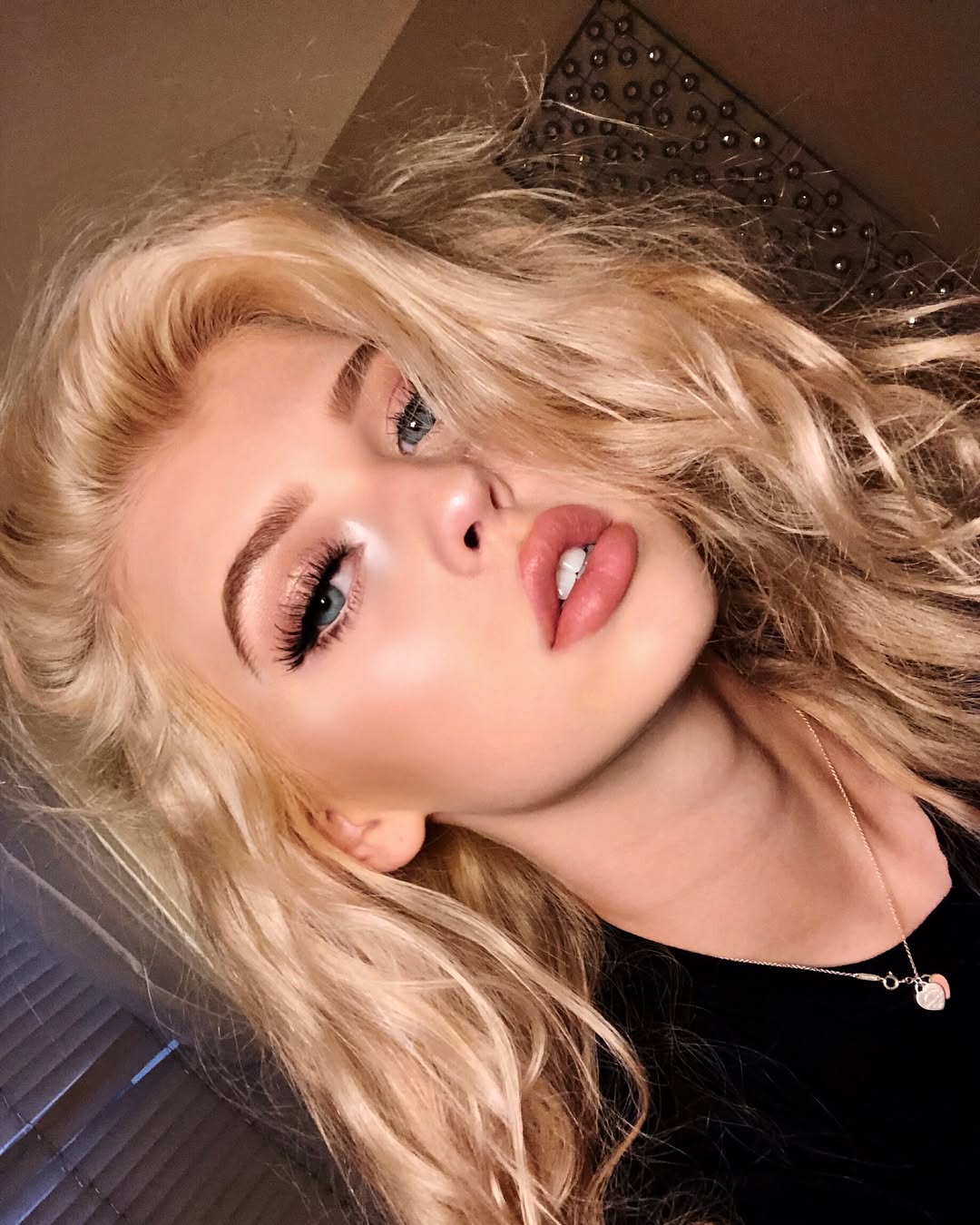 a closeup portrait of a blonde woman with blue eyes wearing makeup and a necklace. she is wearing a black shirt and is posing for the camera.