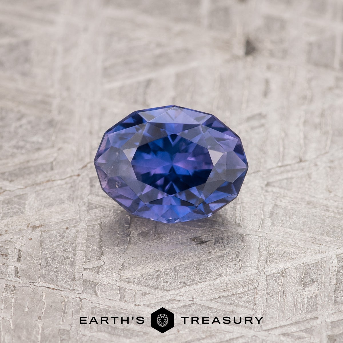 a deep blue sapphire gemstone with an octagonal cut sits on a textured gray surface. the gem is polished and reflects light, showcasing its facets and clarity. the background suggests a stone or metallic texture. the image includes the text "earth's treasury" and a logo featuring a gemlike geometric shape.