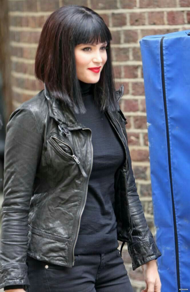 a young woman with long black hair and bangs wears a black leather jacket, black shirt, and red lipstick as she poses against a brick wall in a city setting.