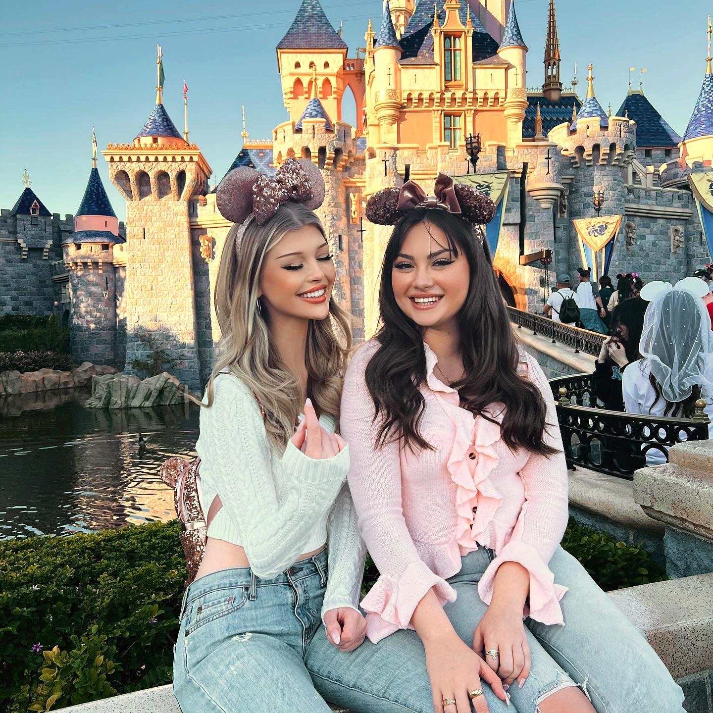 two women are posing in front of disneyland's sleeping beauty castle. they are both wearing mickey mouse ears headbands, jeans, and sweaters. the woman on the left has blonde hair and a white sweater, while the woman on the right has brown hair and a pink sweater. they are both smiling and wearing rings on their fingers. behind them is the iconic castle, a body of water, and other tourists.