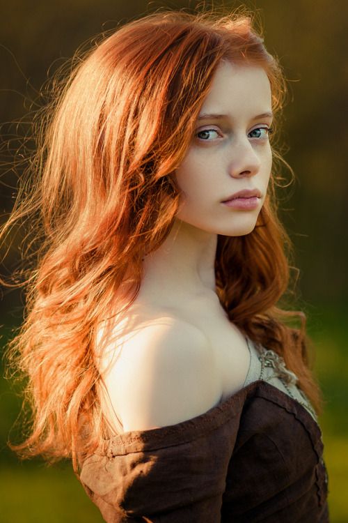 a portrait of a young woman with auburn hair, pale skin, and blue eyes. she is wearing a brown medieval dress that is off the shoulder and is looking directly at the viewer.