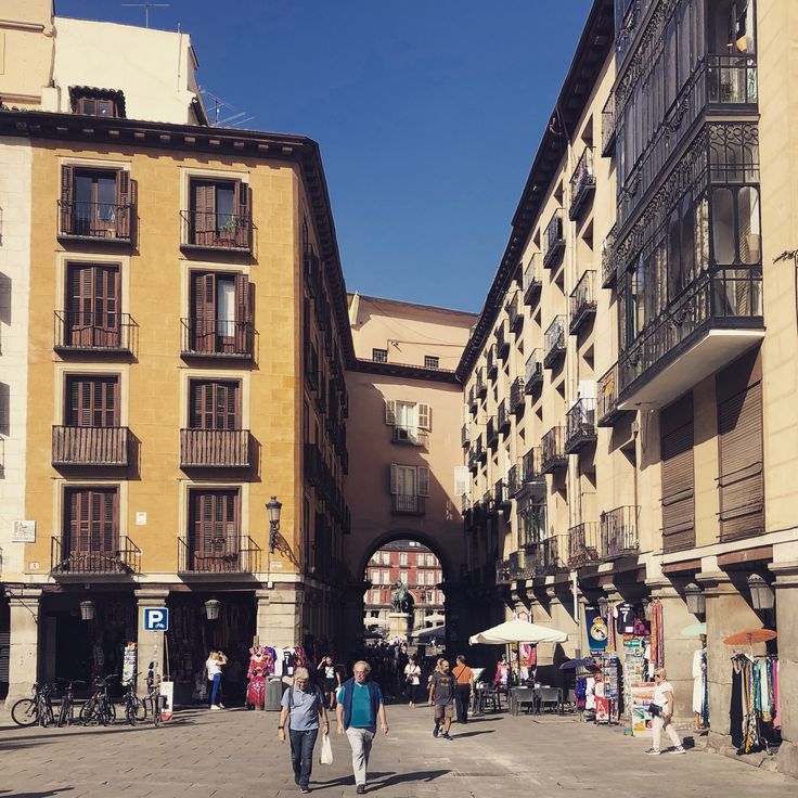 the image shows a sunny street scene in what appears to be the plaza mayor area of madrid, spain.  two rows of tall, historical buildings, primarily yellow and beige, line a narrow street. many of the buildings feature traditional spanish balconies.  an archway is visible in the distance, leading to another open space. the street is populated with pedestrians, some appearing to be tourists, and there are small shops selling souvenirs along the street.  the overall atmosphere is vibrant and characteristic of a historical european city center. the scene is captured from a streetlevel perspective on a bright, clear day.