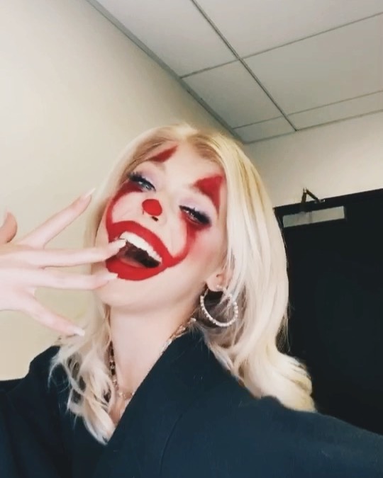a woman with blonde hair is posing for a selfie while wearing clown makeup, which includes red lipstick, red triangles above her eyes, and a red circle around her mouth and nose. she is wearing large hoop earrings, a necklace, and a black jacket. she has her fingers near her mouth and is looking at the camera with a playful expression. the background appears to be a room with beige walls and a dark door.