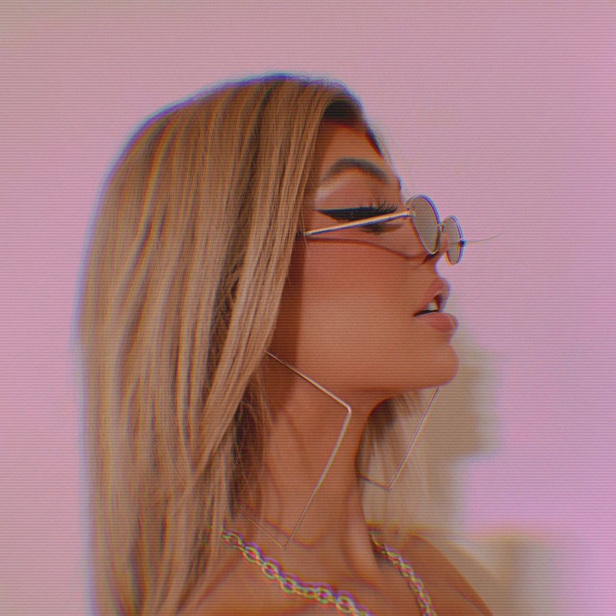a profile shot of a woman with blonde hair wearing sunglasses, square earrings, and a chain necklace against a pink background with a shadow. the woman has eyeliner on.